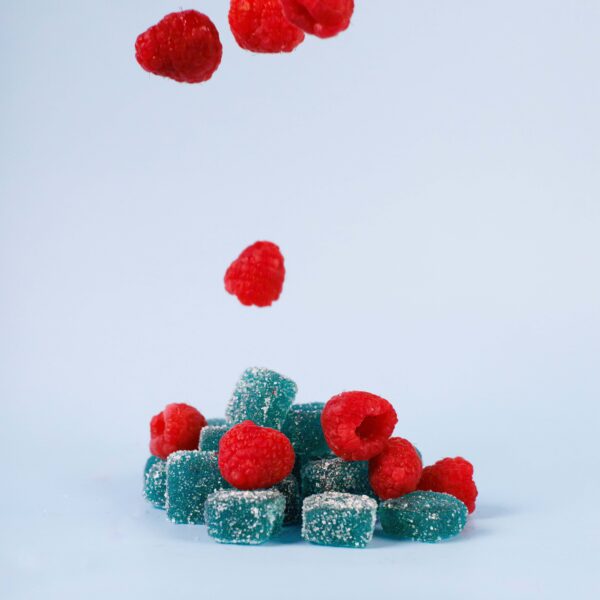Raspberries and blue gummy candies falling and stacked, with sugar coating, on a light blue background, representing tasty and colorful confectionery products, inspired by modern confectionery design.
