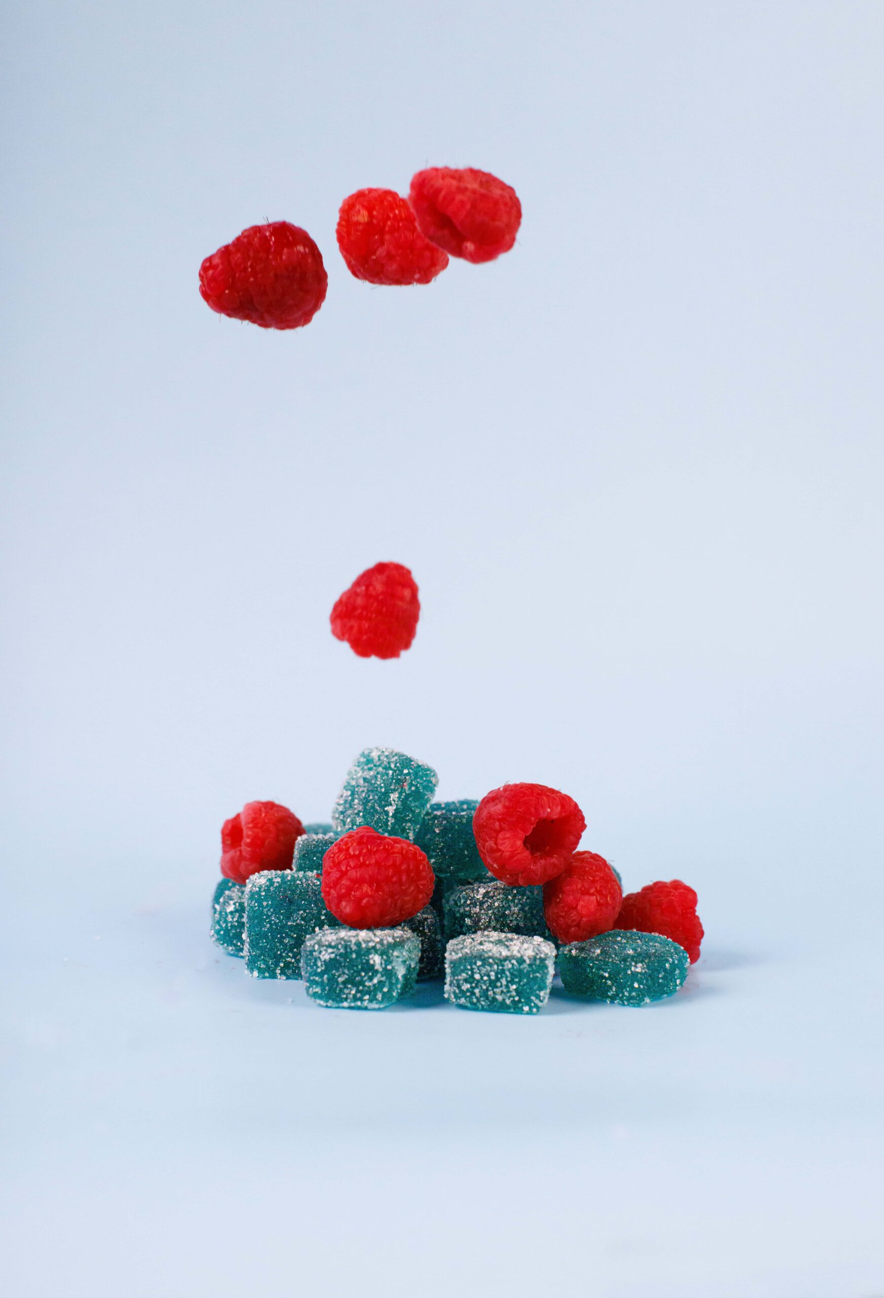 Raspberries and blue gummy candies falling and stacked, with sugar coating, on a light blue background, representing tasty and colorful confectionery products, inspired by modern confectionery design.